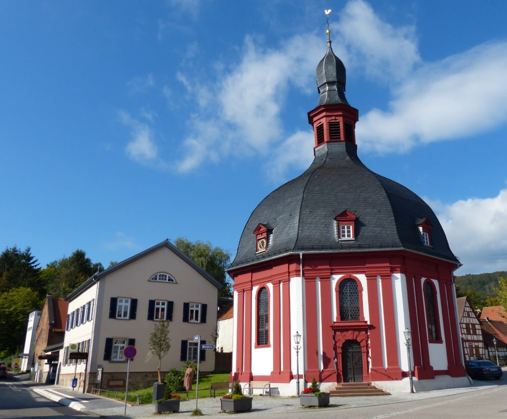 Außenbild von Kirche mit Pfarrhaus vor einem blauen Himmel mit Wolken. 