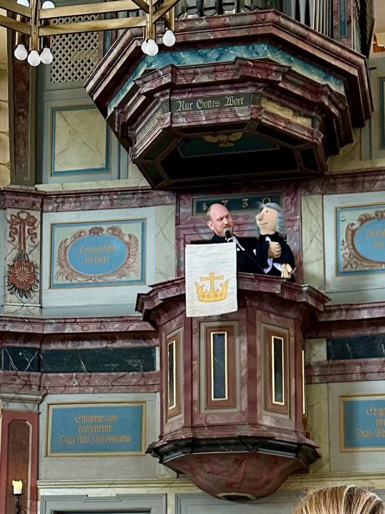 Pfarrer Ebling steht mit der Puppenfigur Pater Valentino auf der Kanzel