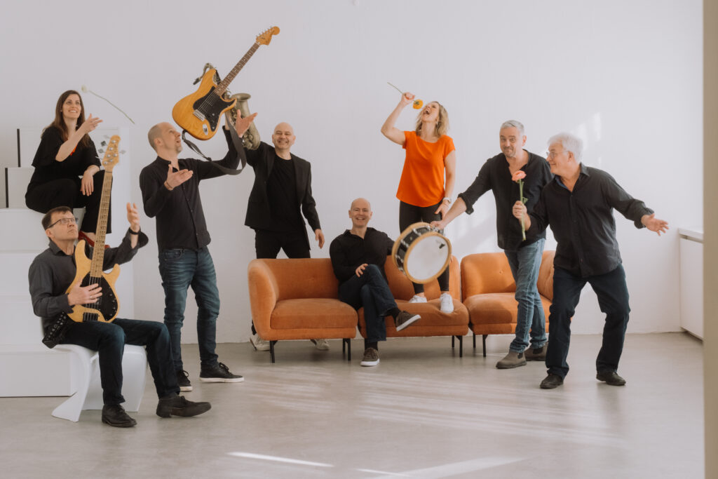 Es sind sechs Männer und zwei Frauen vor einer weißen Wand zu sehen,die zum Teil auf einem Sofa sitzen und zum Teil stehen. Manche haben ein Instrument oder eine Blume in der Hand. Es ist ein Bild mit lockerer Stimmung. 