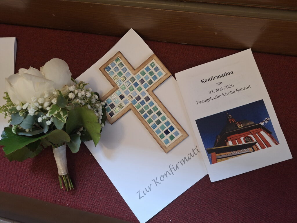 Auf einer Kirchenbank liegt ein Blumensträußchen, eine Urkunde, ein selbstgestaltetes Kreuz mit Mosaiksteinen sowie das Liedblatt. 