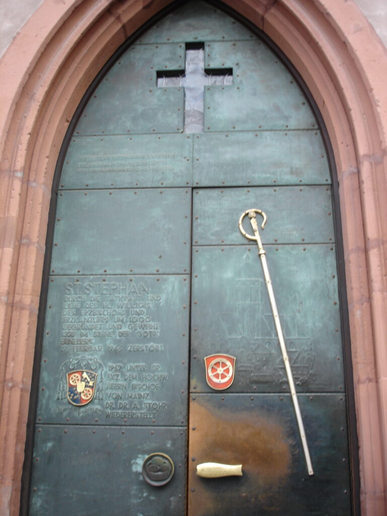 Bild zeigt das Eingangsportal der Kirche - eine schwere, schwarze Tür mit Beschriftung, einem ausgeschnittenen Kreuz, zwei Wappen und einem Stab. Umrahmt ist die Tür von Sandstein.