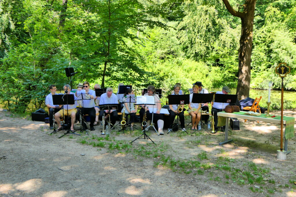 Blasorchester sitzt, im Hintergrund sind Bäume und der Weiher