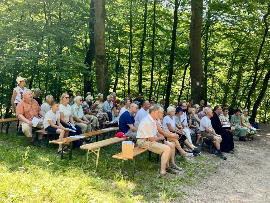 Die Gottesdienstbesucher sitzen auf Bierbänken vor grünen Bäumen.
