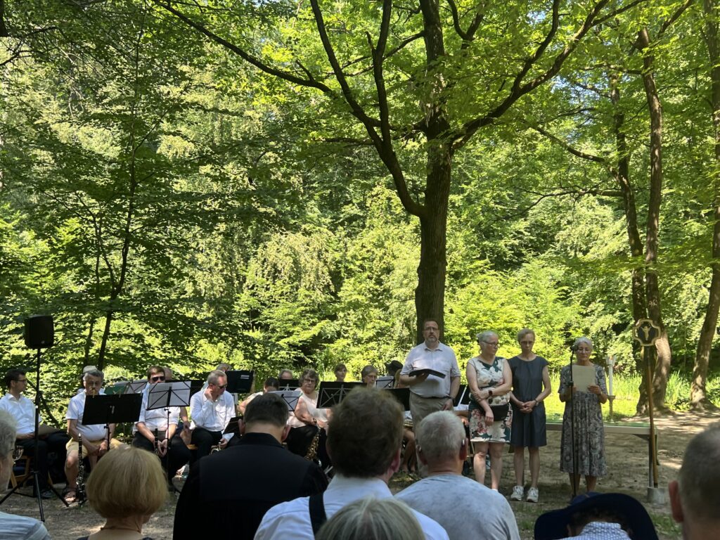 Es sind Köpfe von Gottesdienstbesuchern von hinten zu sehen und dahinter linbks sitzend Mitglieder des Blasorchesters und rechts sdtehend vier Personen, die im Gottesdienst mitgewirkt haben.