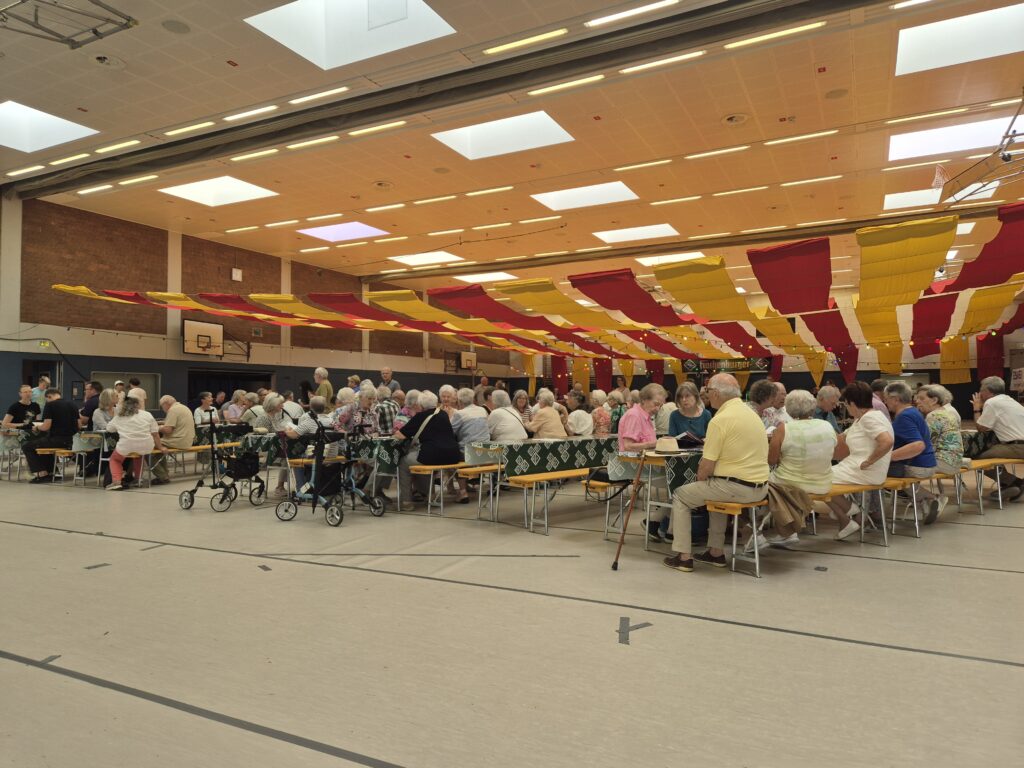 Viele Menschen sitzen in Reihe an Bierzeltgarnituren in der Sporthalle.