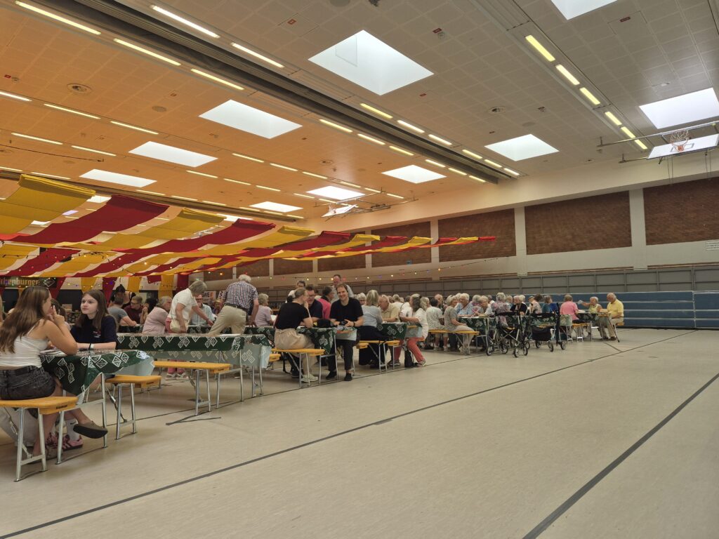 Viele Menschen sitzen in Reihe an Bierzeltgarnituren in der Sporthalle.