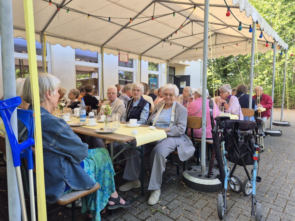 Menschen sitzen an Tischen und Bänken vor dem Gemeindehaus