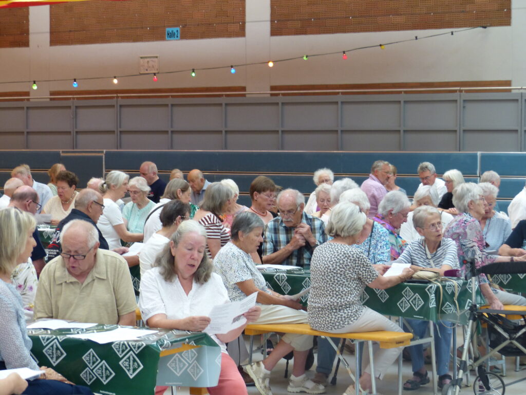 Viele Menschen sitzen in Reihe an Bierzeltgarnituren in der Sporthalle.