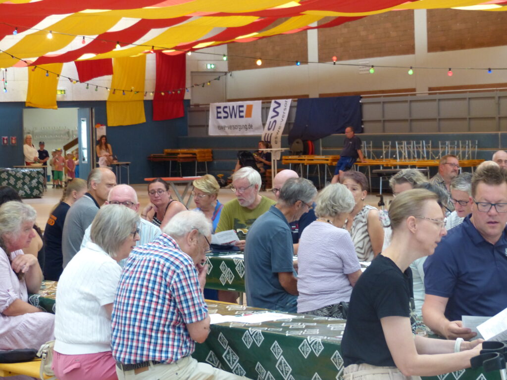Viele Menschen sitzen in Reihe an Bierzeltgarnituren in der Sporthalle.