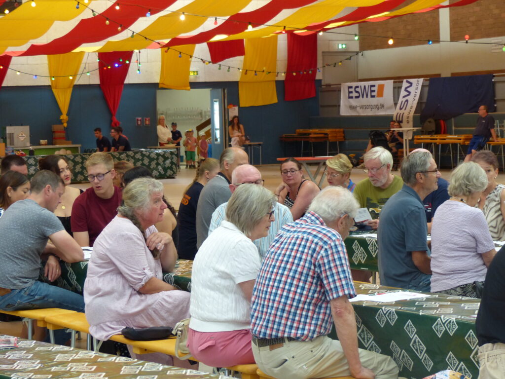 Viele Menschen sitzen in Reihe an Bierzeltgarnituren in der Sporthalle.