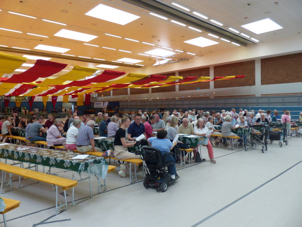 Viele Menschen sitzen in Reihe an Bierzeltgarnituren in der Sporthalle.