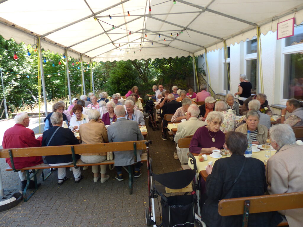 Menschen sitzen an Tischen und Bänken vor dem Gemeindehaus