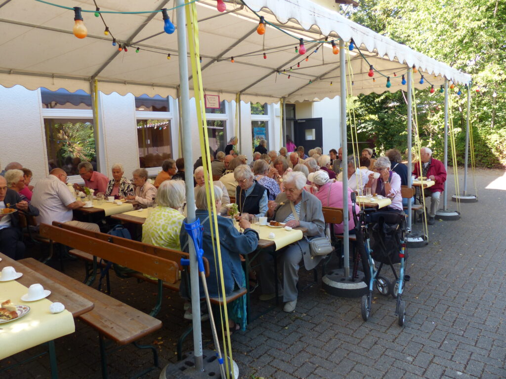 Menschen sitzen an Tischen und Bänken vor dem Gemeindehaus