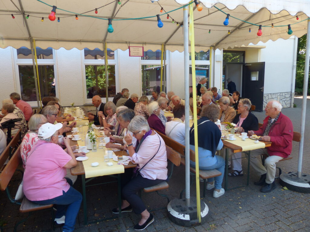 Menschen sitzen an Tischen und Bänken vor dem Gemeindehaus